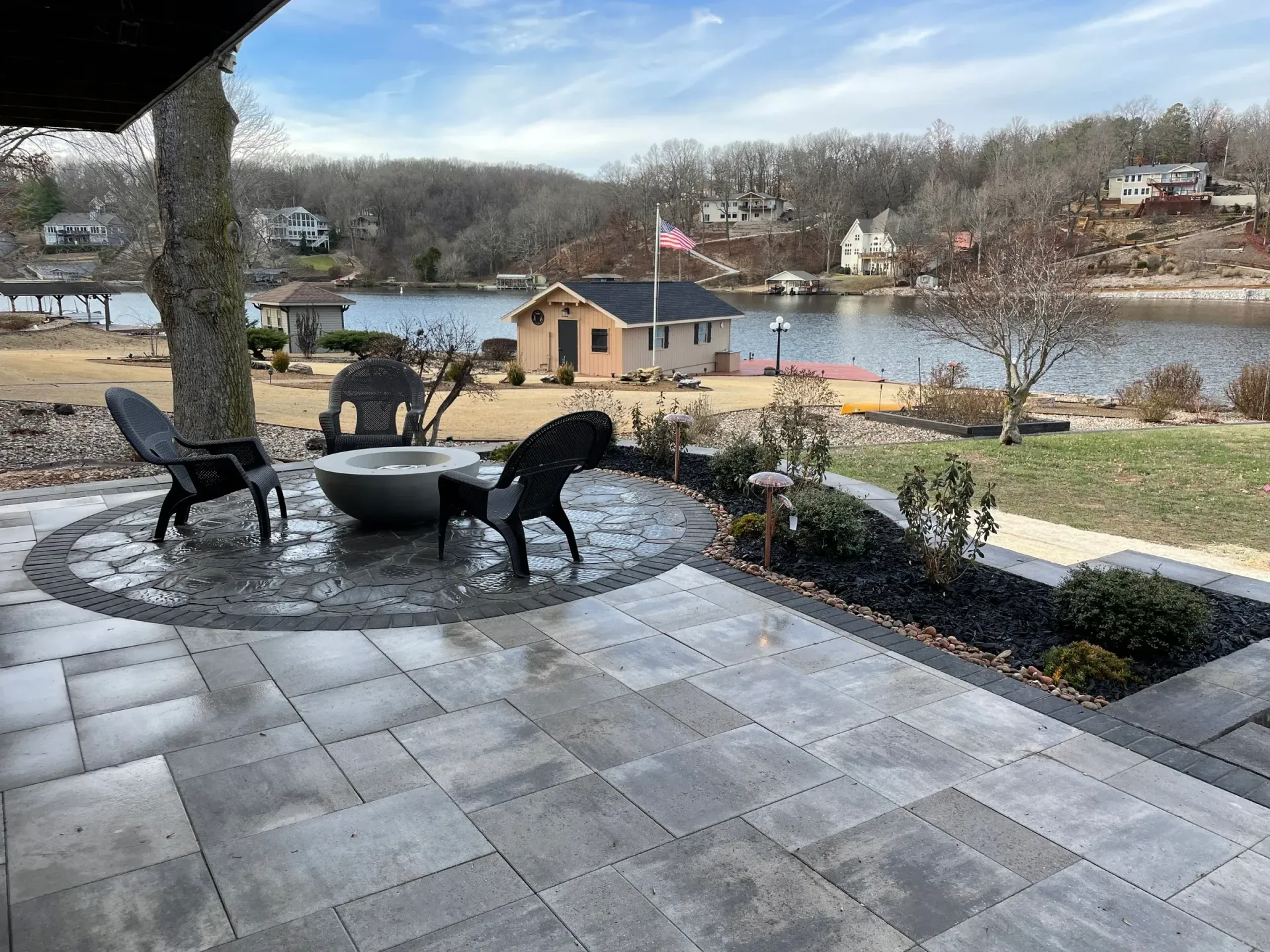 Patio overlooking a lake with two chairs by a fire pit. Distant houses and trees.