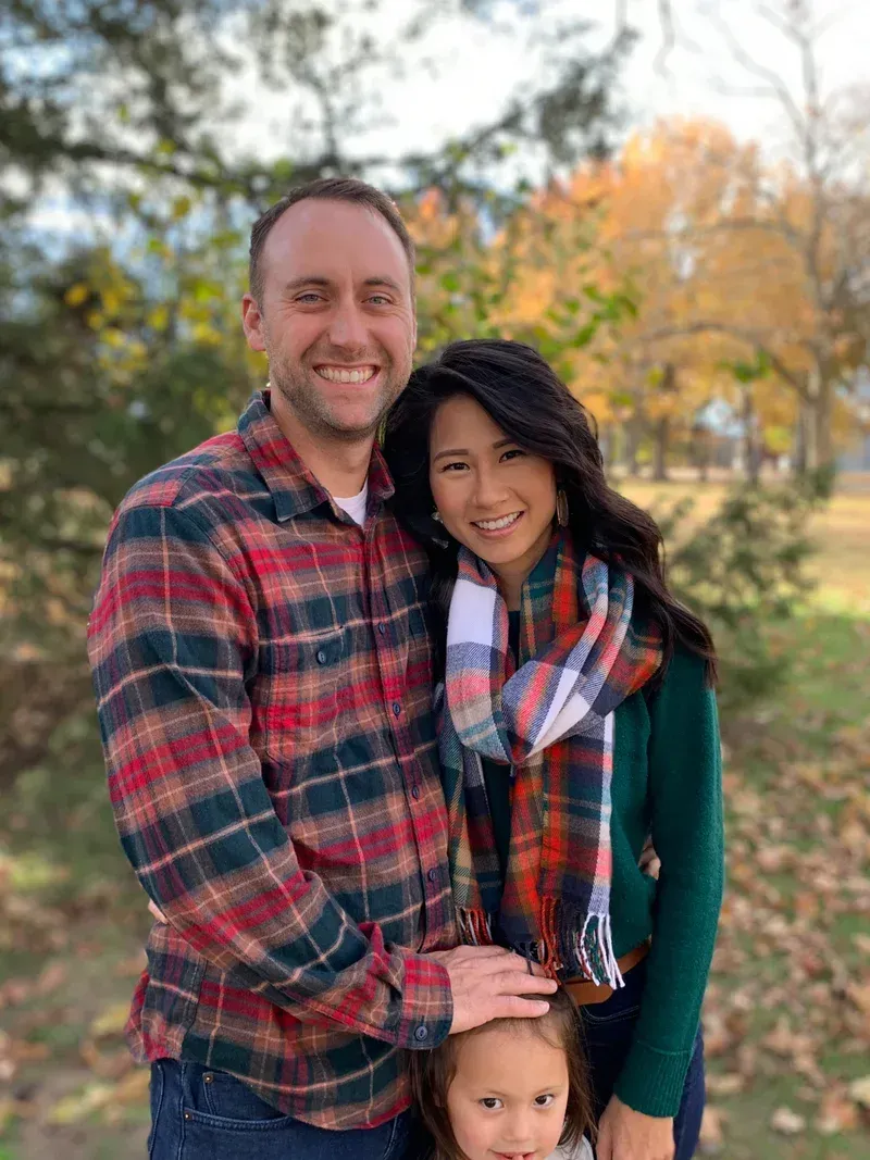 Smiling couple with a child pose outdoors. They wear fall colors: plaid shirts, green sweater, scarf.