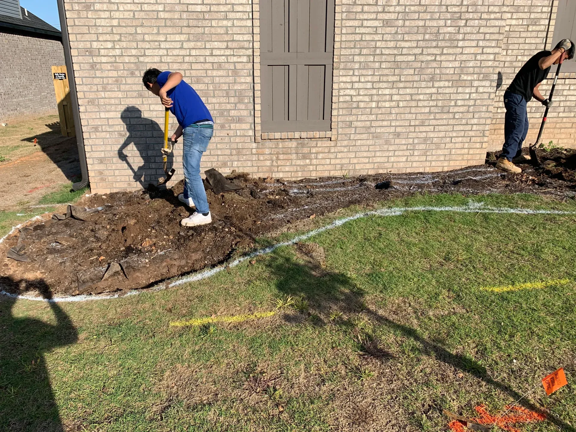 Two people digging in a garden bed next to a brick building.