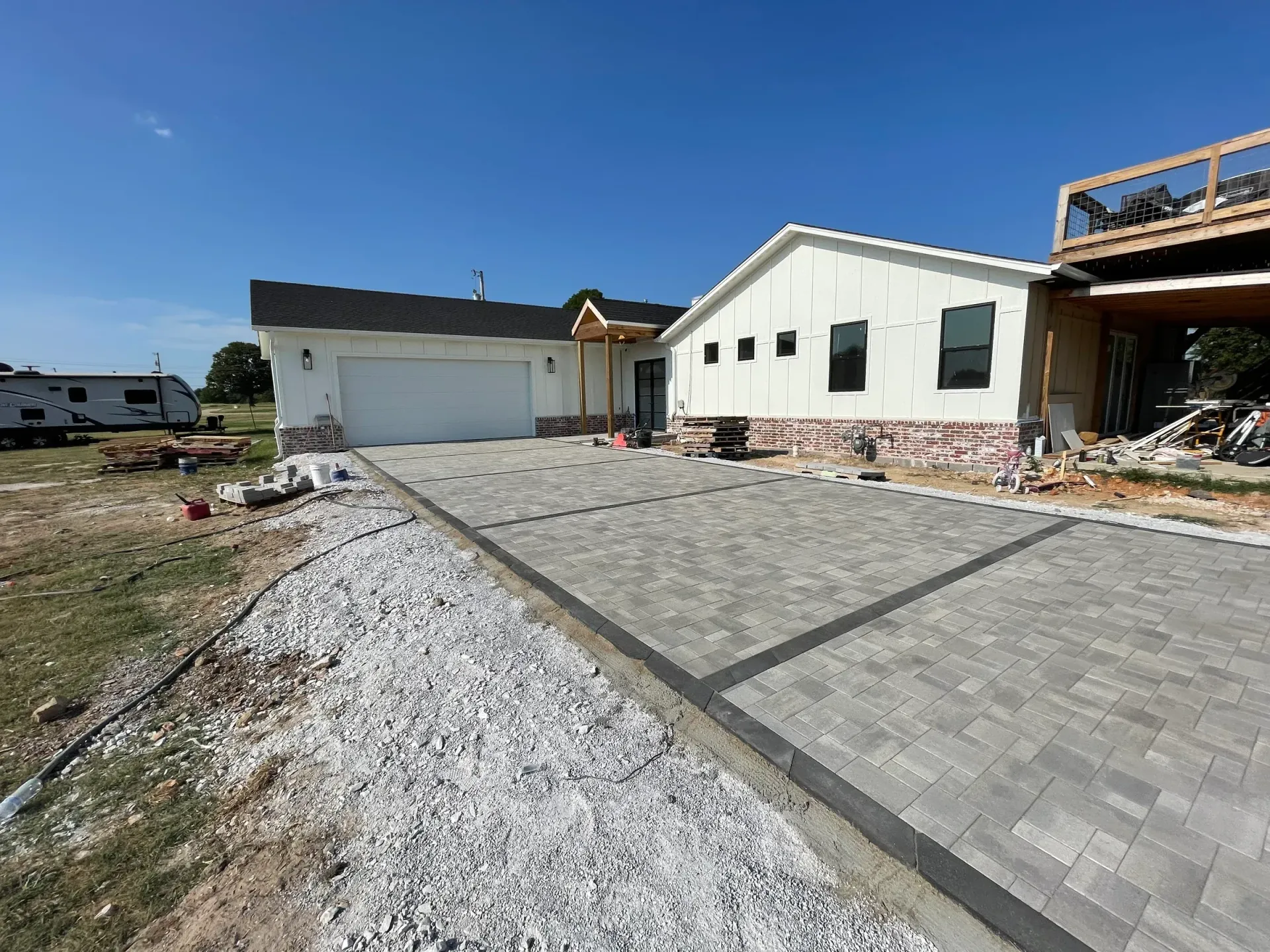 Driveway with brick pavers leading to a white house with a garage and new construction deck, under a clear blue sky.