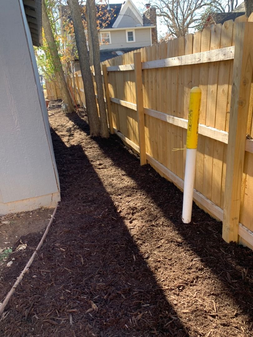Wood fence and mulch path beside a building. Yellow post. Trees.