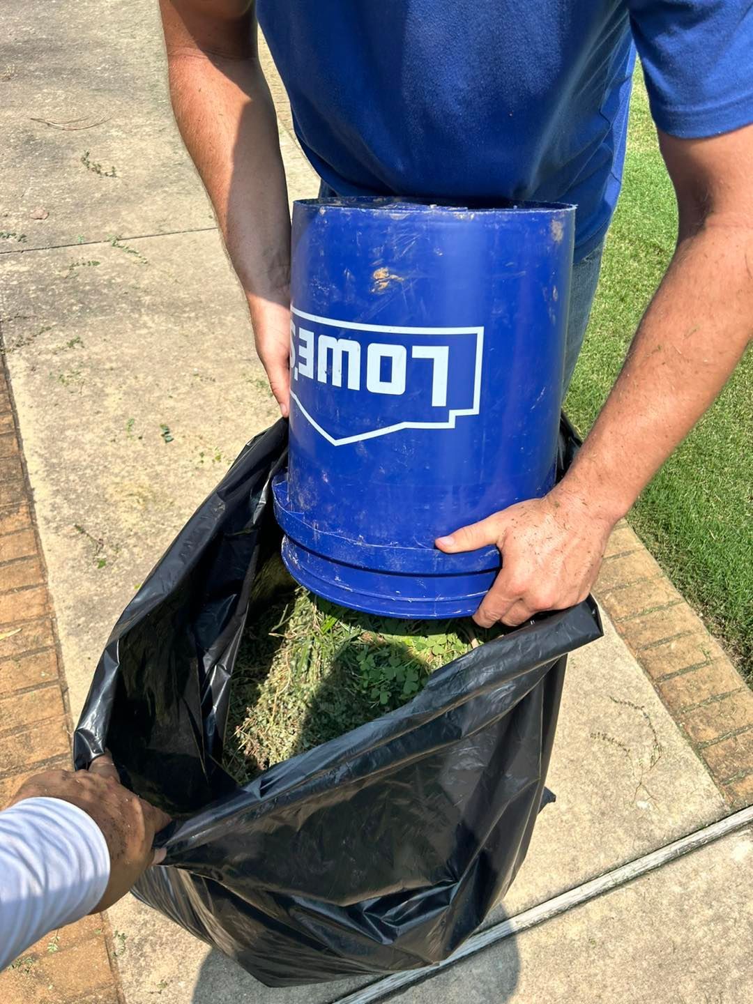 Person emptying grass clippings from a blue Lowe's bucket into a black trash bag on a concrete surface.