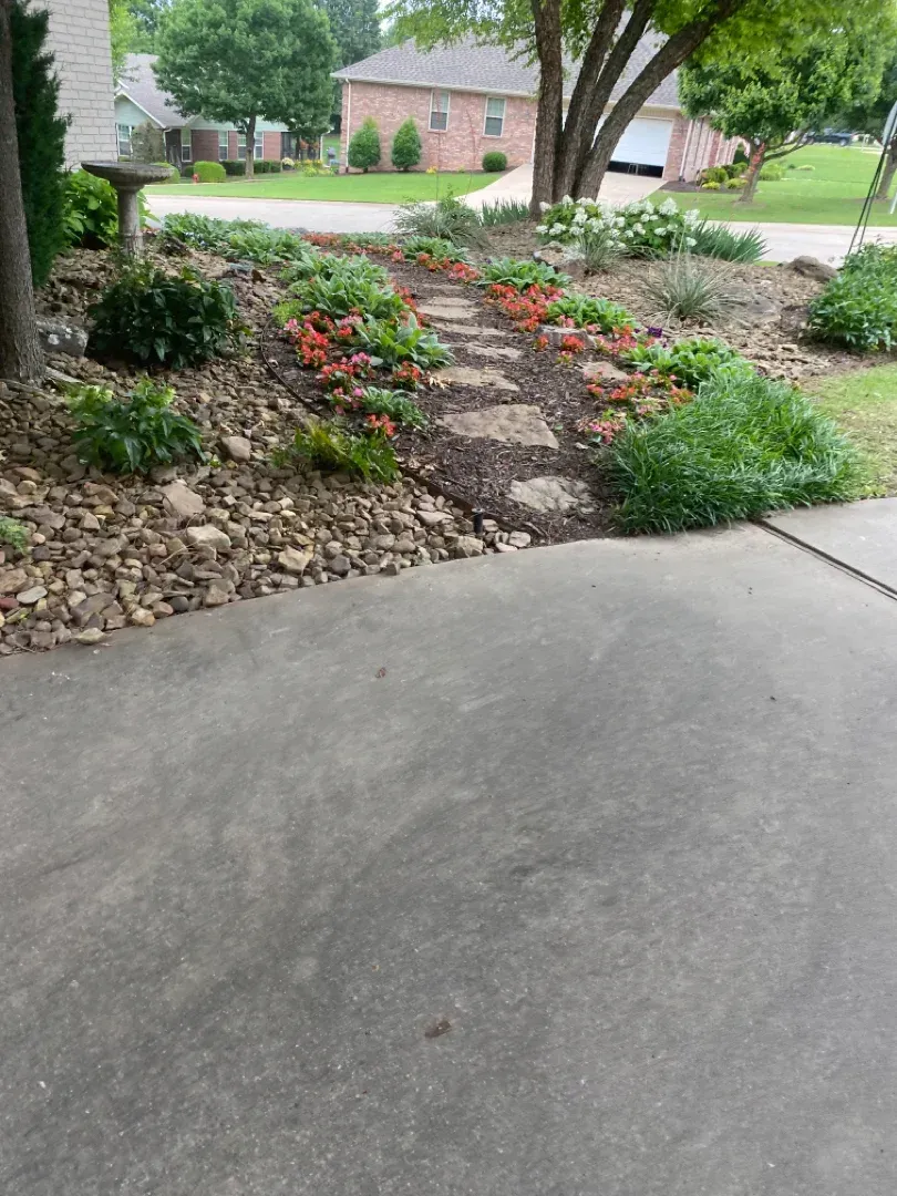 Stone path leads through a landscaped garden with colorful flowers, set in a front yard, by a house.