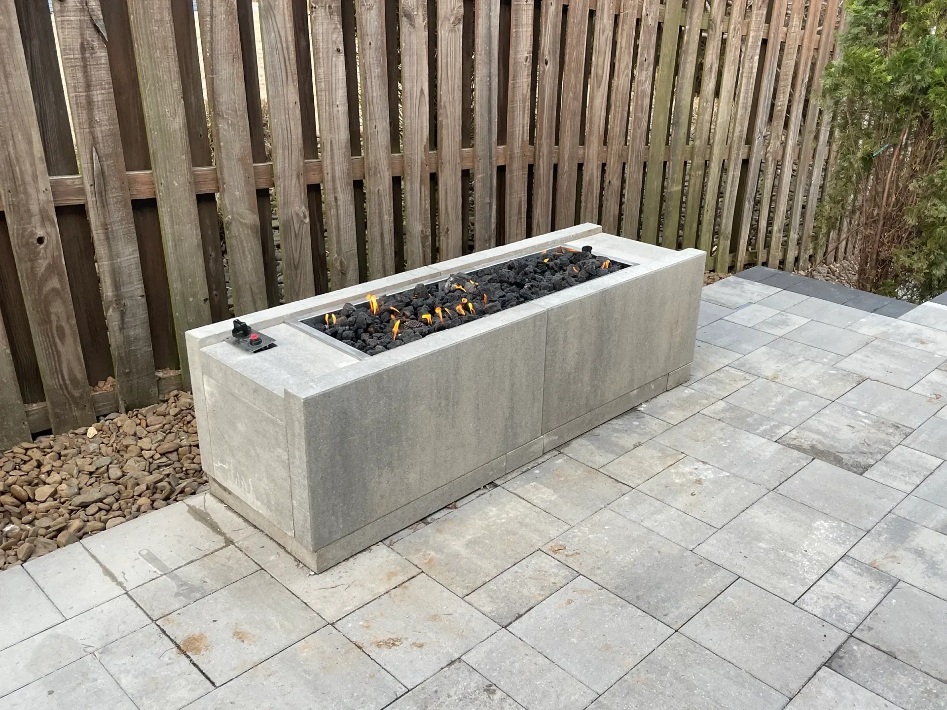 Rectangular concrete fire pit with burning flames on a paved patio, near a wooden fence.
