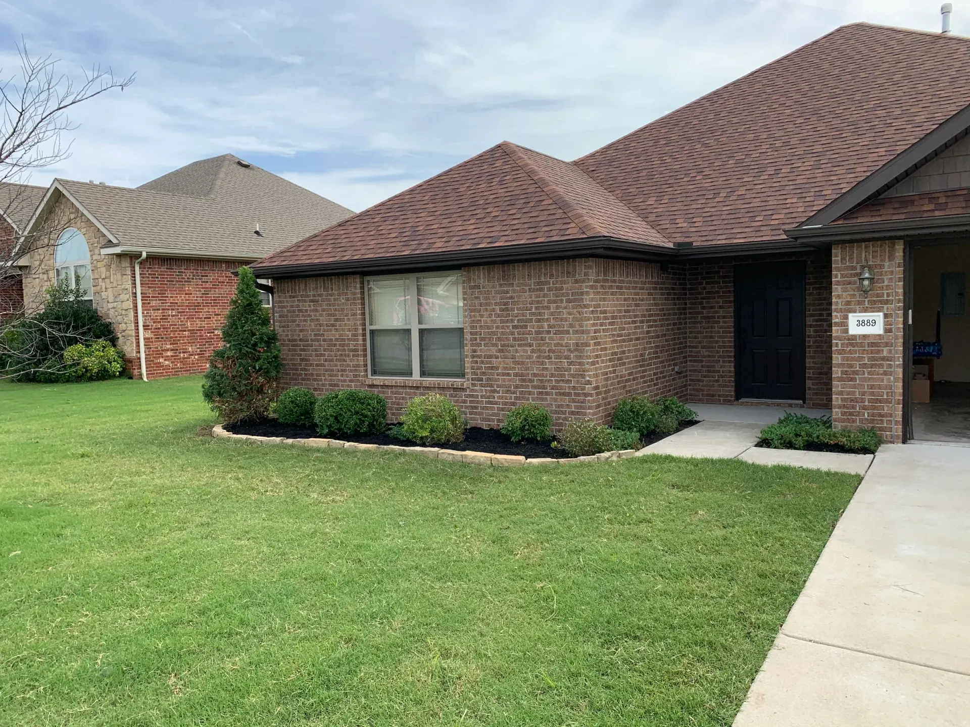 Brick house with brown roof, green lawn, landscaping, and sidewalk.