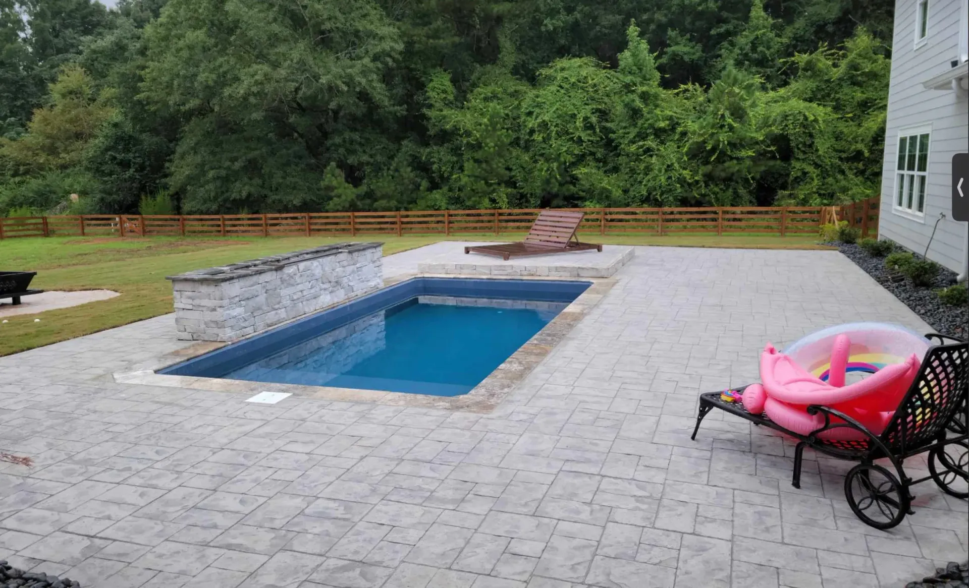 Backyard with rectangular pool, patio, and lounge chairs; greenery and fence in the background.