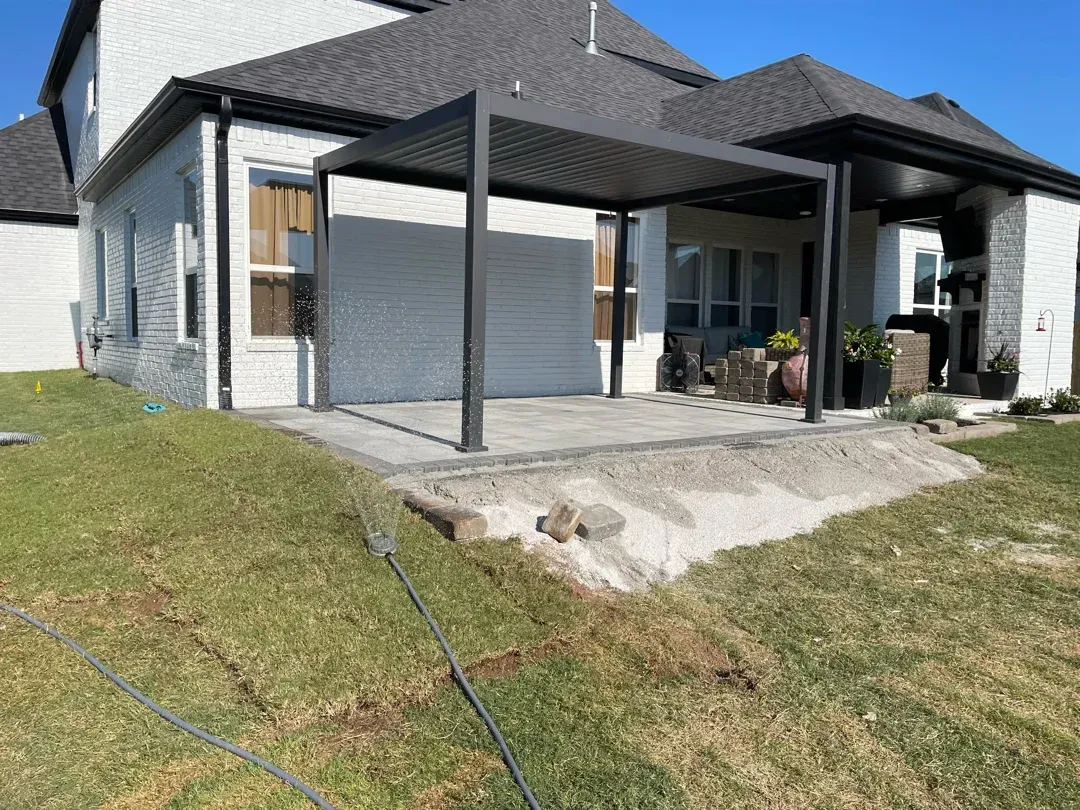 Backyard patio with pergola, gray pavers, white brick house, and green grass.