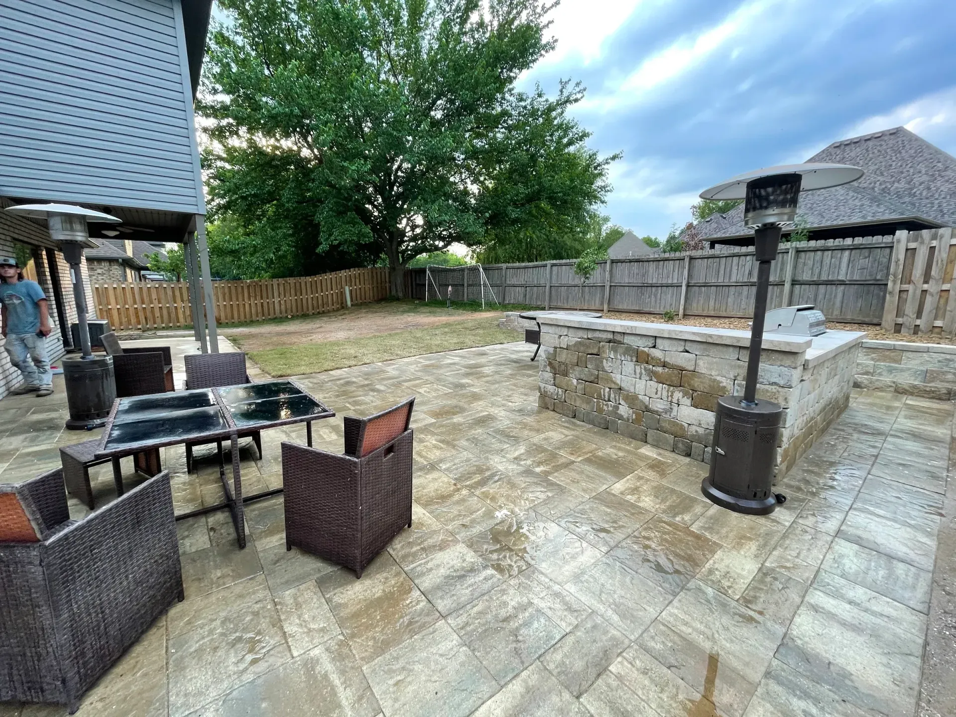 Patio with outdoor kitchen, seating, and heaters. Stone pavers, wooden fence, cloudy sky.