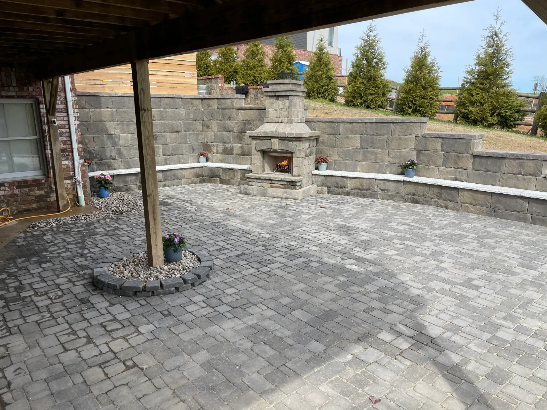 Stone patio with fireplace and retaining wall. Underneath a wooden deck, small trees in background.