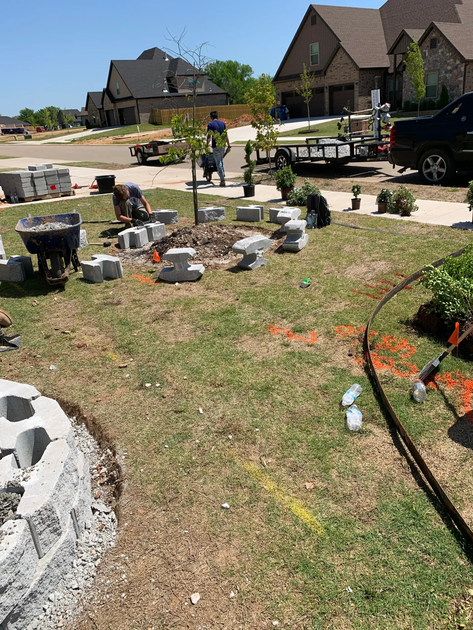 Workers building a stone border around a tree in a residential yard. Blocks, tools, and materials scattered around.