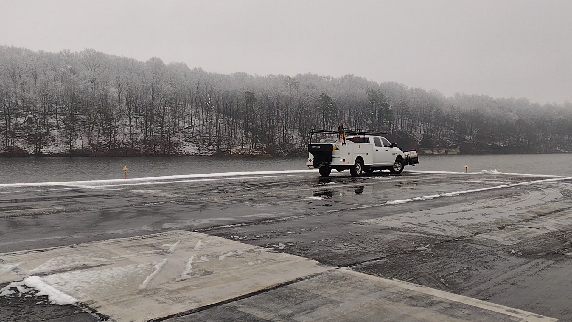 White truck parked on a snowy boat ramp. Trees and water in background under overcast sky.