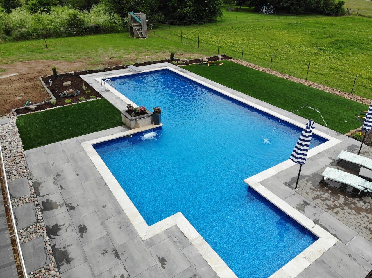Rectangular blue tiled swimming pool with light-colored coping, surrounded by paved patio and green grass.