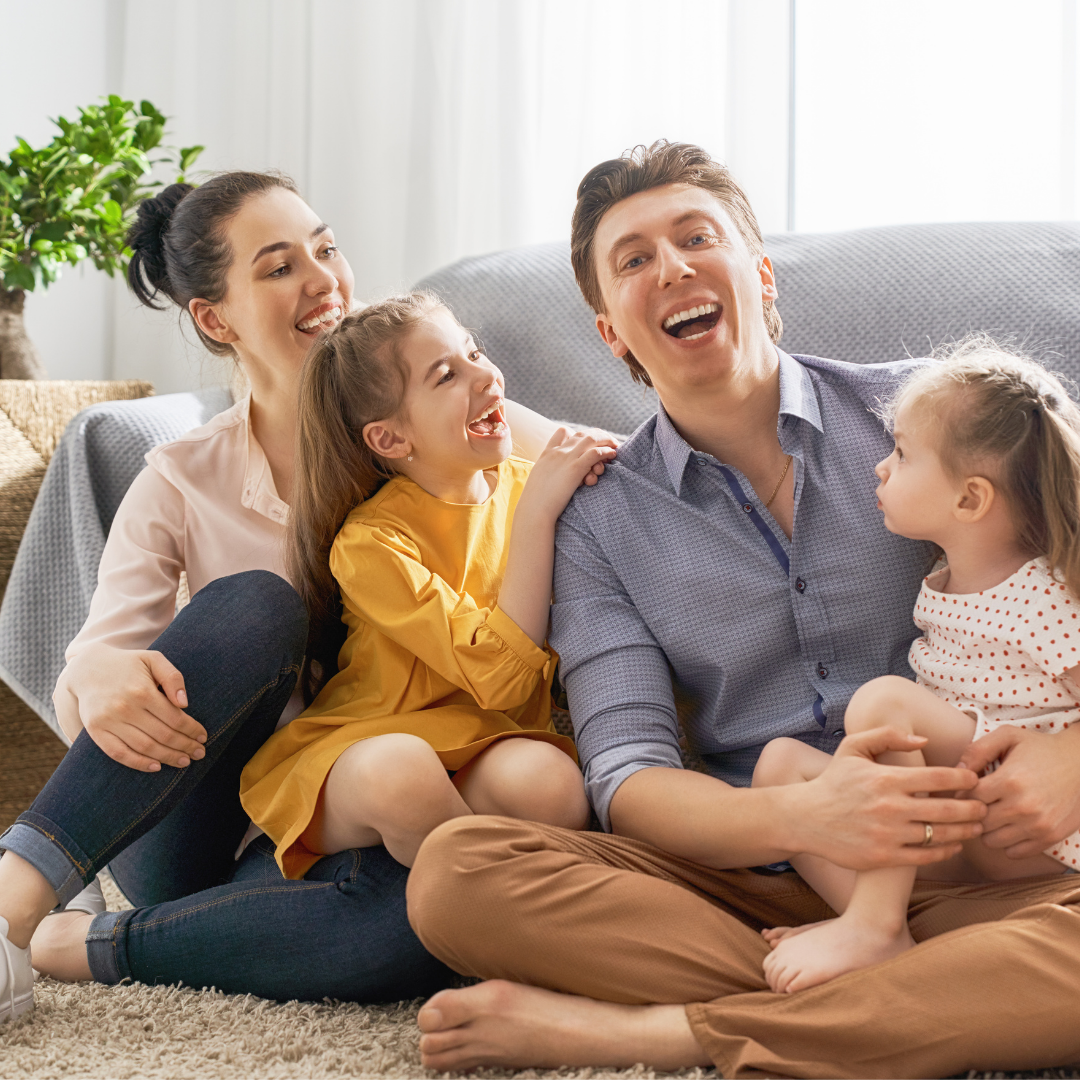 Family laughing together indoors: mother, father, and two young children sitting on the floor.