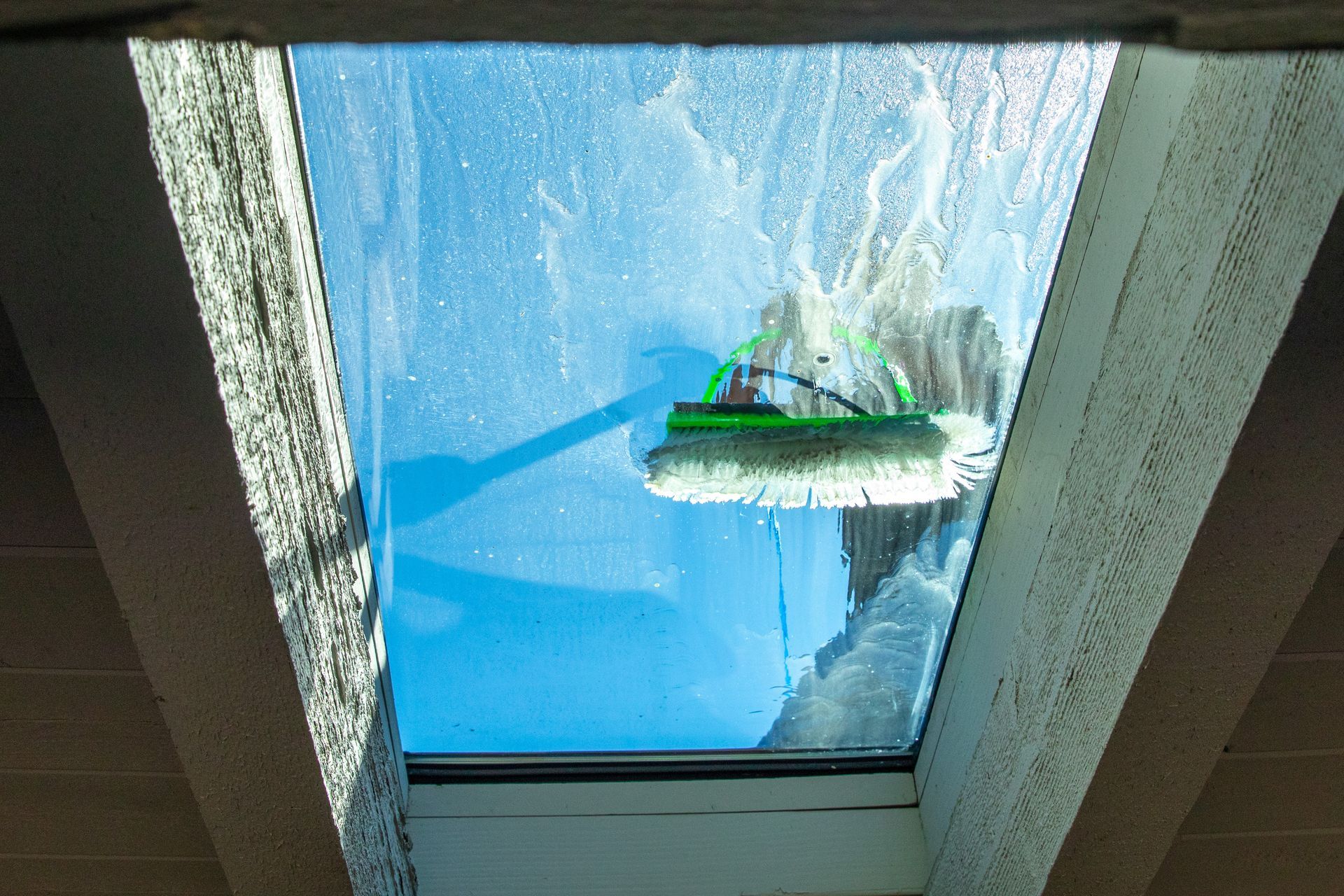 A green-bristled brush on an extension pole cleans a skylight window, revealing a blue sky above.