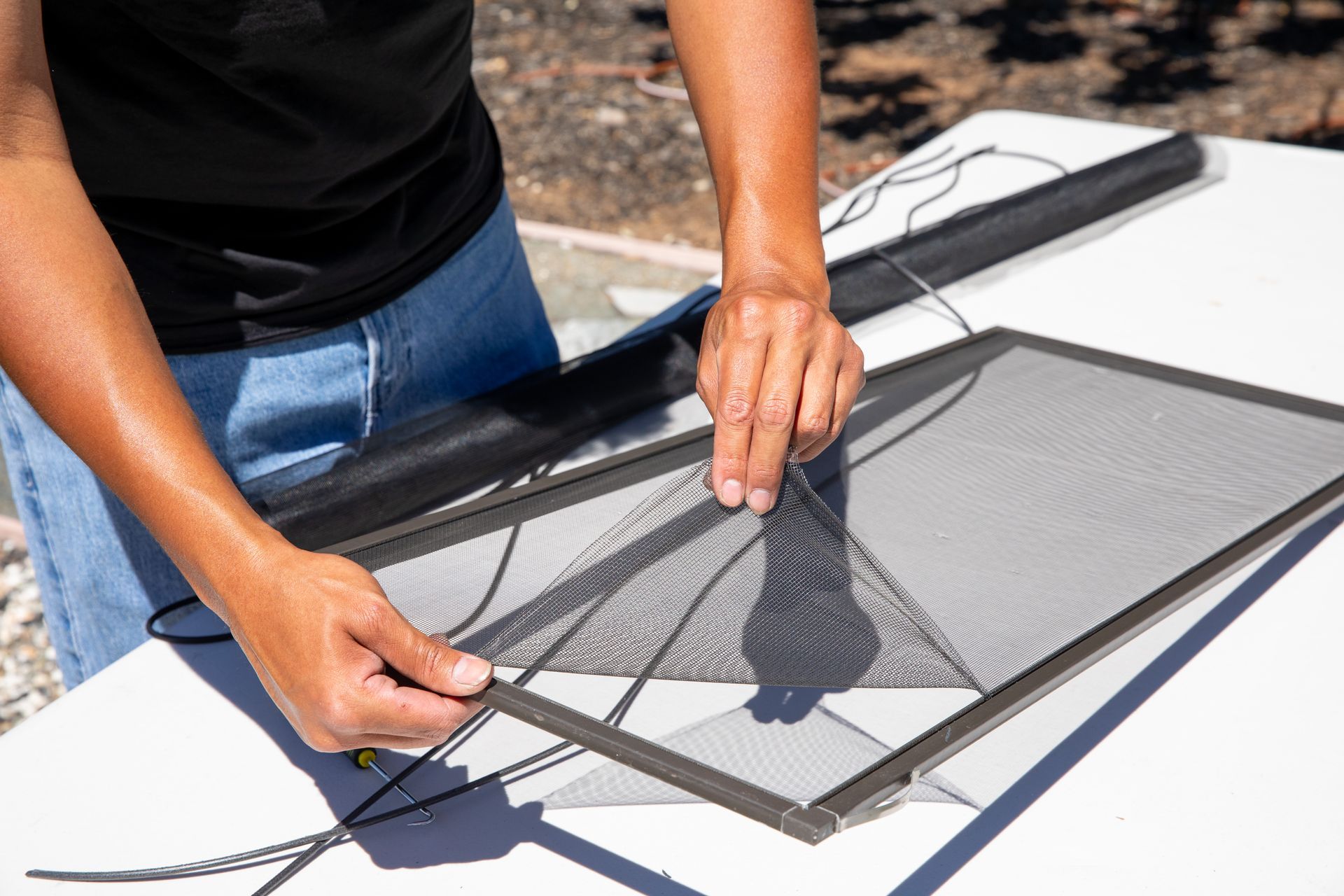 Hands secure black mesh screen material into the metal frame of a window screen using a spline tool on a white table.