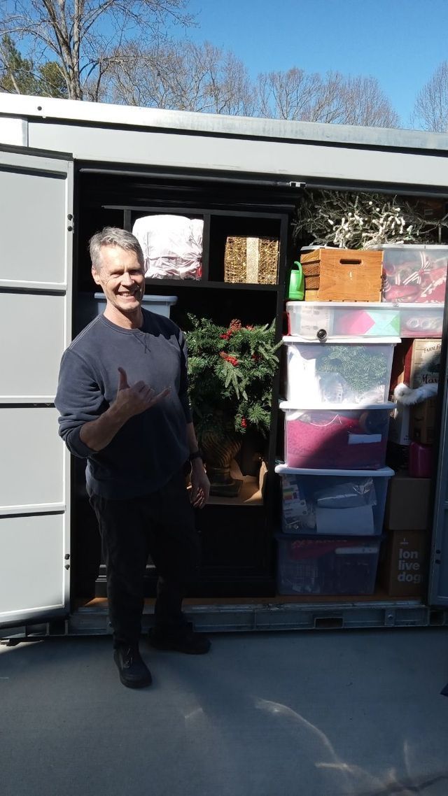 Person stands beside stacked storage bins outside a moving truck, waving and smiling.