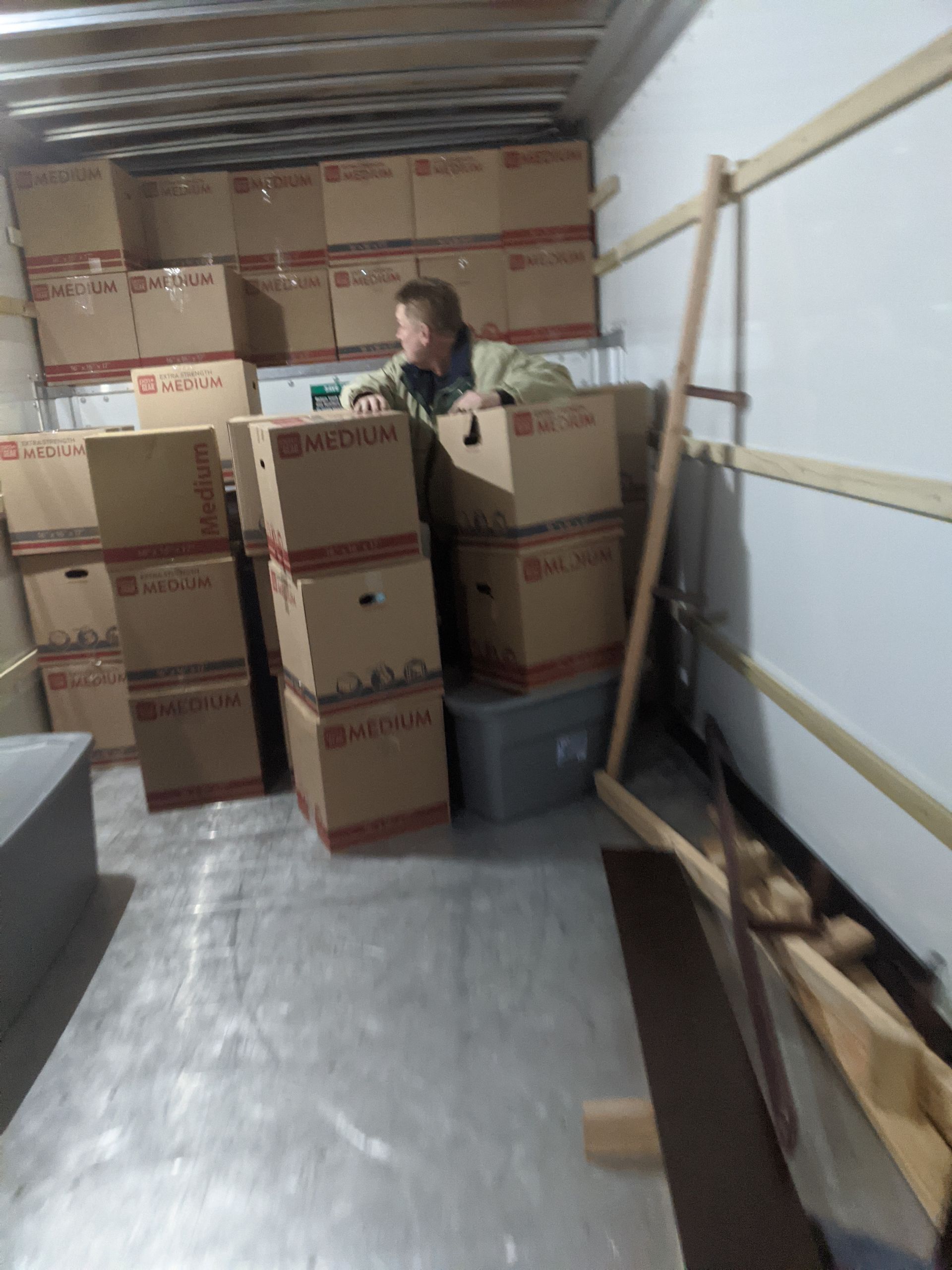 Person standing among stacked cardboard boxes in a narrow storage area, with a ladder on the right.