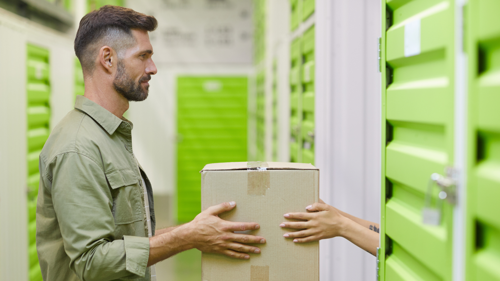 A person hands a cardboard box to another person inside a self-storage facility with rows of lime-green metal doors.