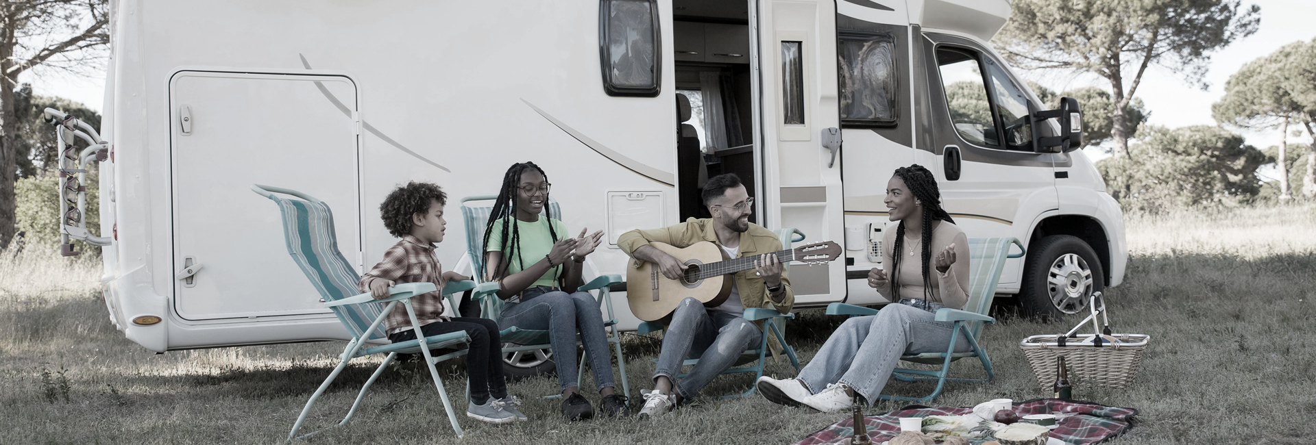 A family sits in chairs by a camper, one person plays guitar, others sing.