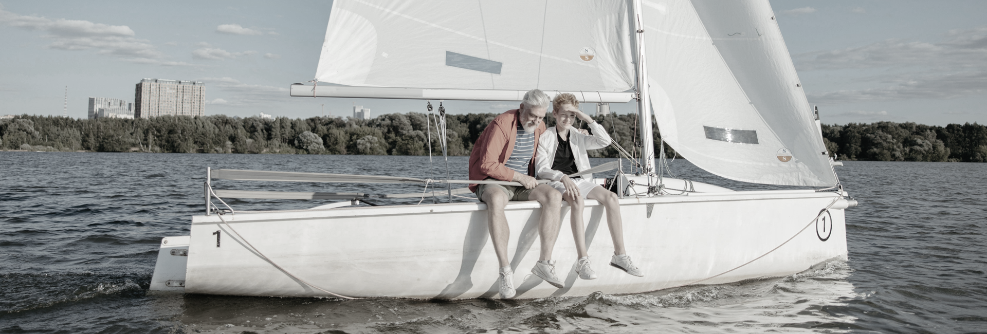 Two people sit on the edge of a sailboat, looking out at a body of water.