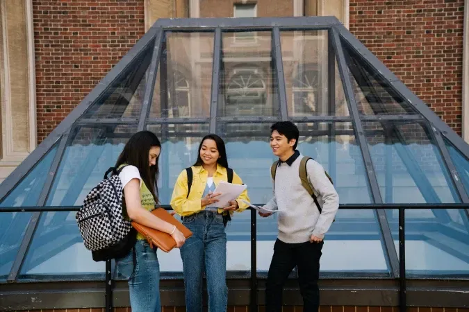 Three students talking on a building's balcony; brick building with glass pyramid roof in background.