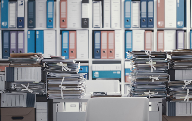 Office desk overwhelmed with stacks of paperwork; file folders line the background shelves.