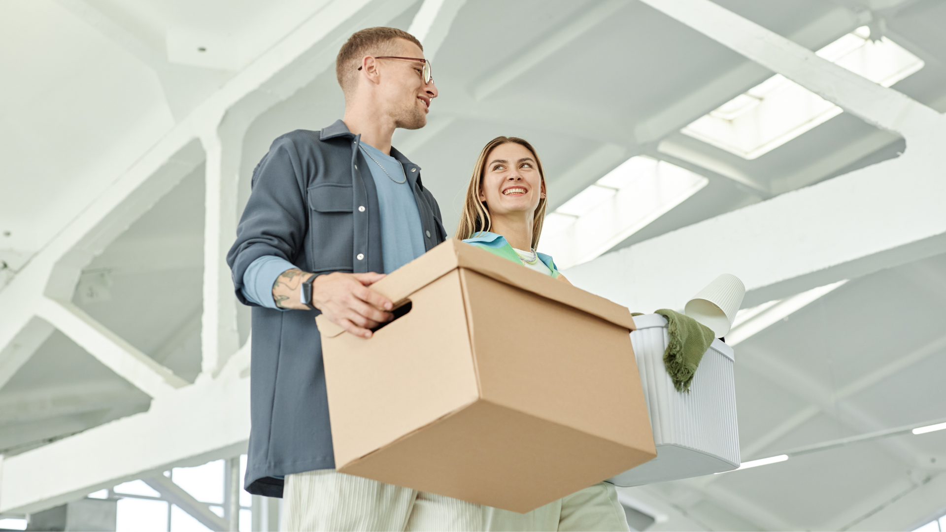 A person in a gray shirt and a person in a turquoise shirt walk together carrying cardboard boxes in a bright room.
