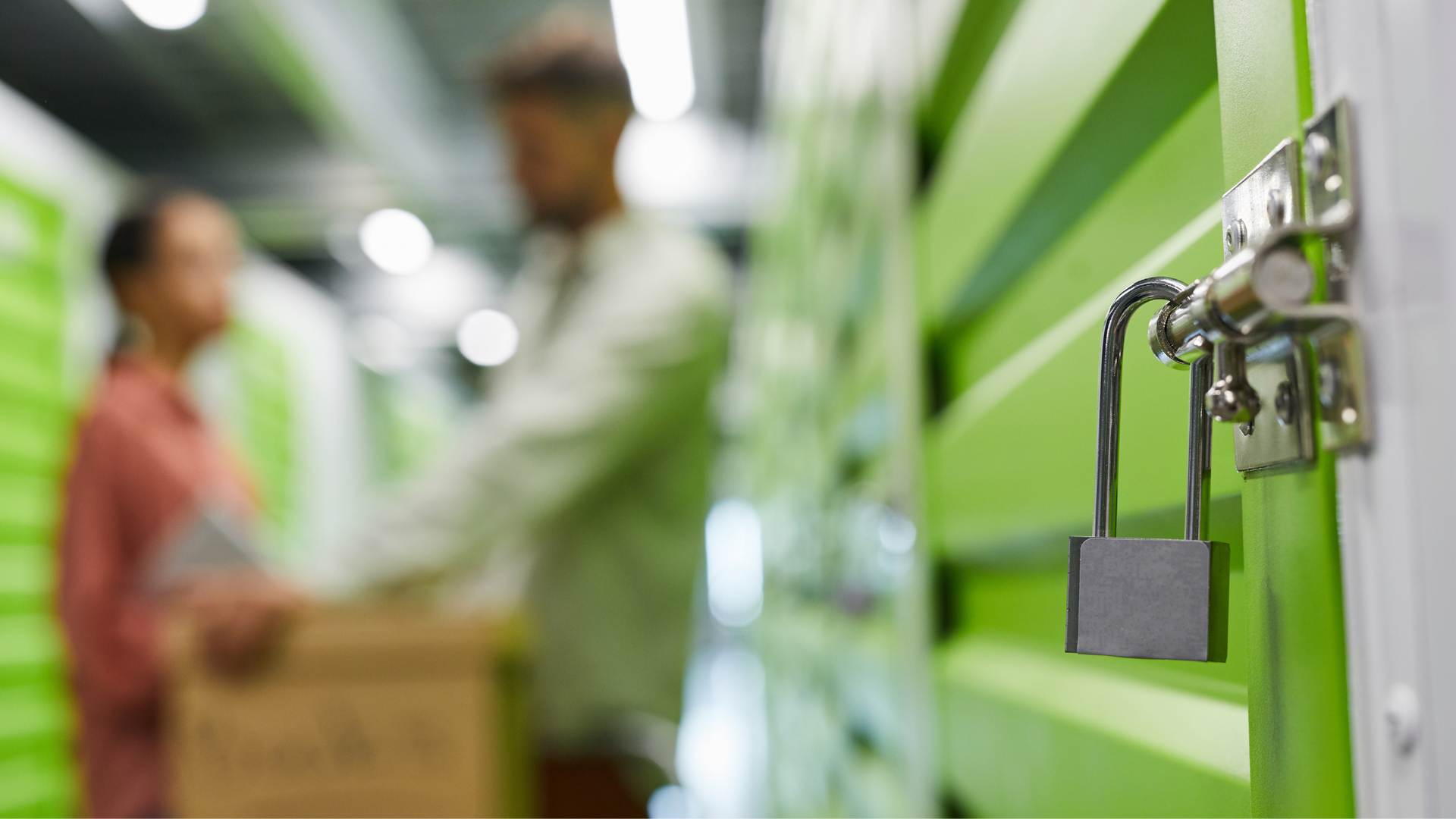 A focus on a silver padlock on a bright green storage unit door, with two blurred people holding boxes in the background.