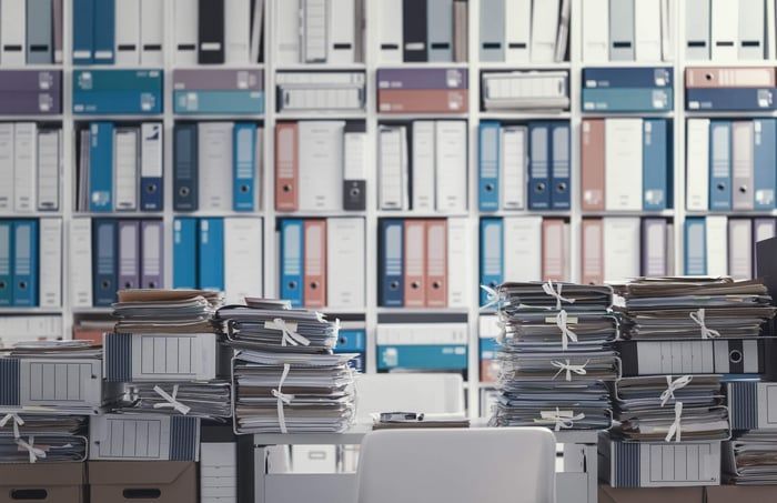 Office desk overwhelmed by piles of paper in front of shelves filled with binders.