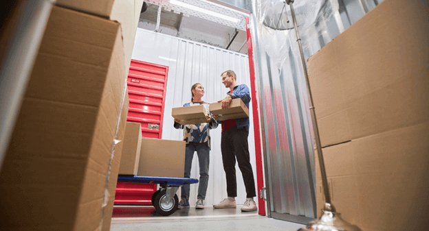Two people loading boxes into a storage unit. Cart and boxes are in the foreground, unit door is red.