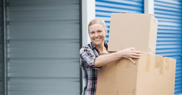 Woman smiling, holding stacked cardboard boxes in front of a blue storage unit.
