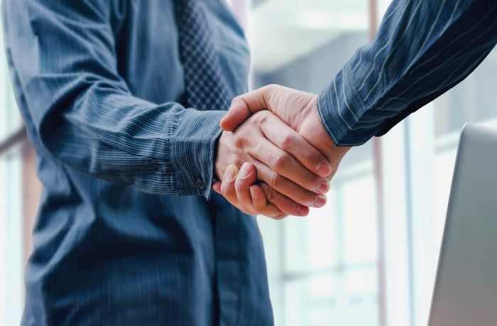 Two people in business attire shaking hands, indoors near a window and laptop.