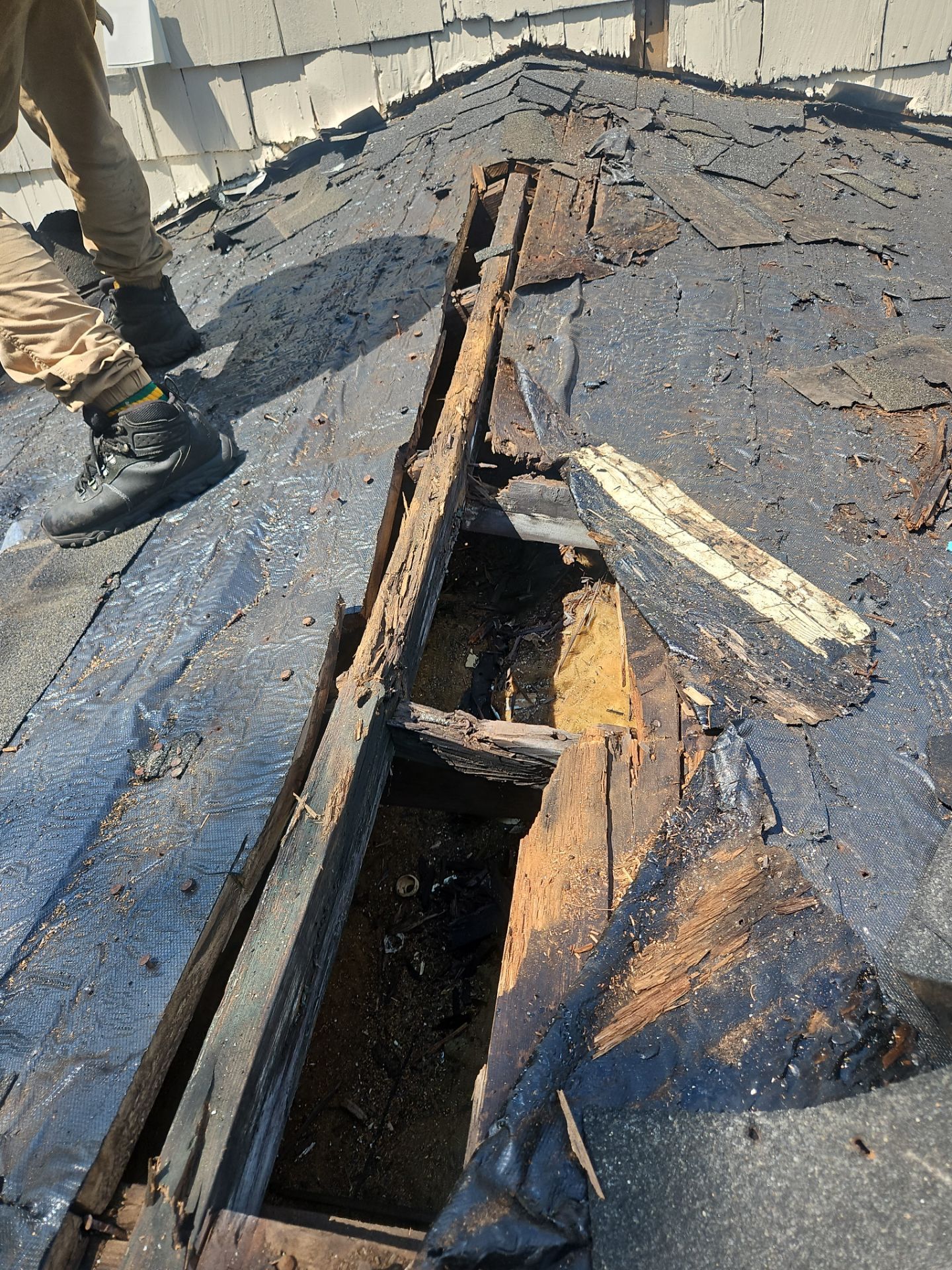 Damaged roof with exposed wood beams and tar paper. Someone wearing boots stands nearby.