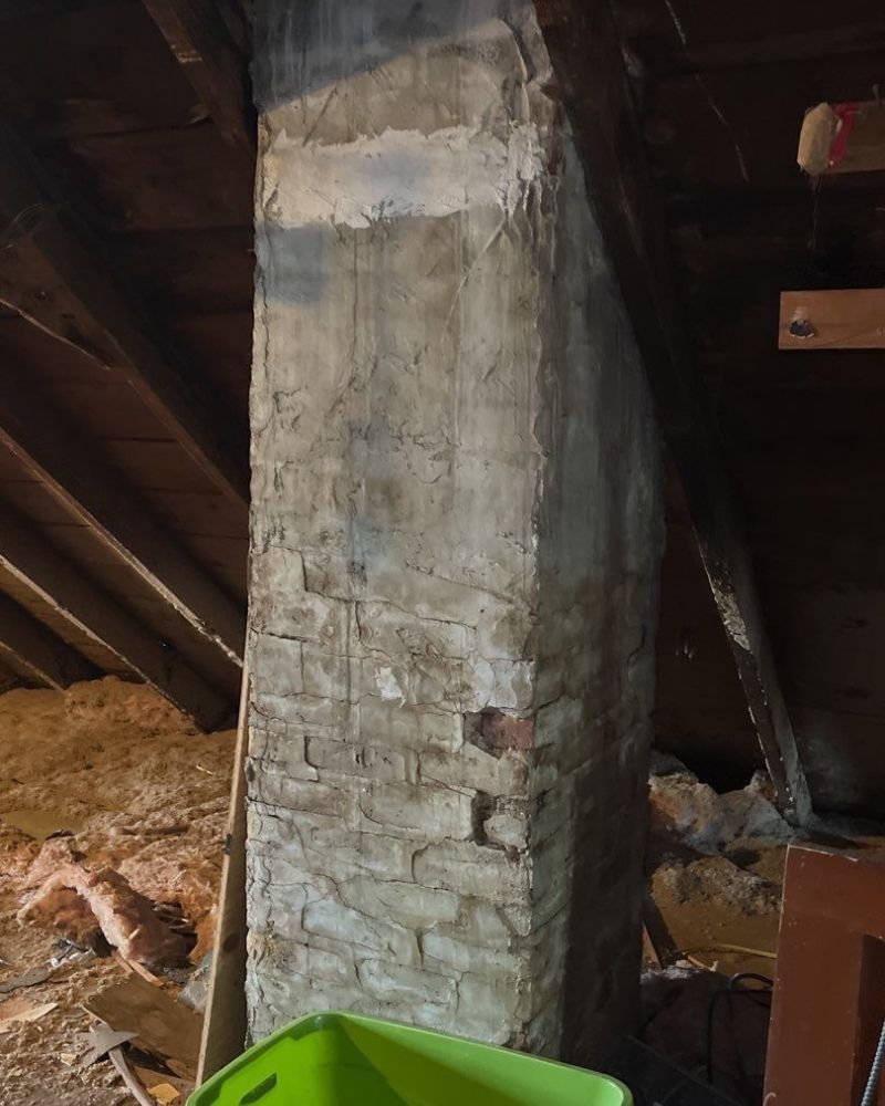 Brick chimney in an attic, with stucco coating in places, framed by wooden rafters and insulation.