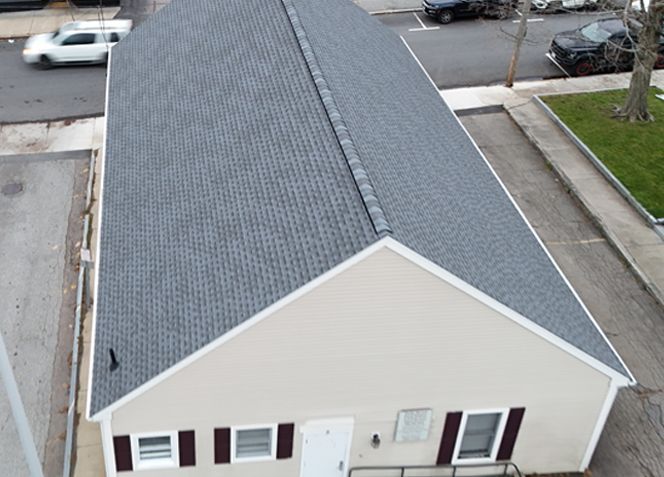 A building with a gray shingle roof and beige siding, seen from above. Black shutters and a white door.