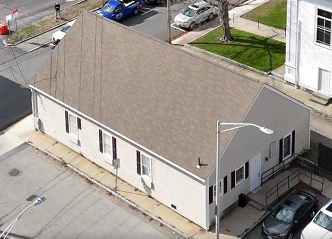 Building with a tan roof and siding, black shutters. Cars and street in view.