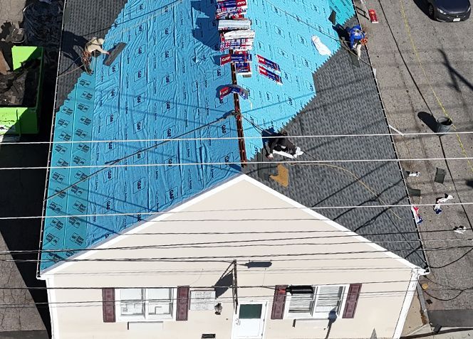 Aerial view of a house roof partially covered with blue underlayment and gray shingles; workers install roofing.