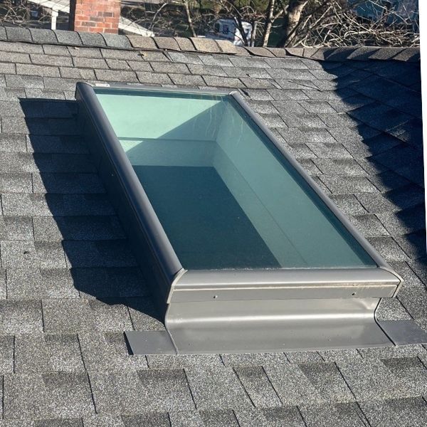 Skylight on a shingled roof, silver frame, clear glass. Sunlight reflects inside the boxy opening.