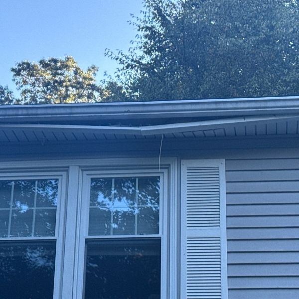 Damaged section of a house gutter. Gray siding and white shutters below. Trees in the background.