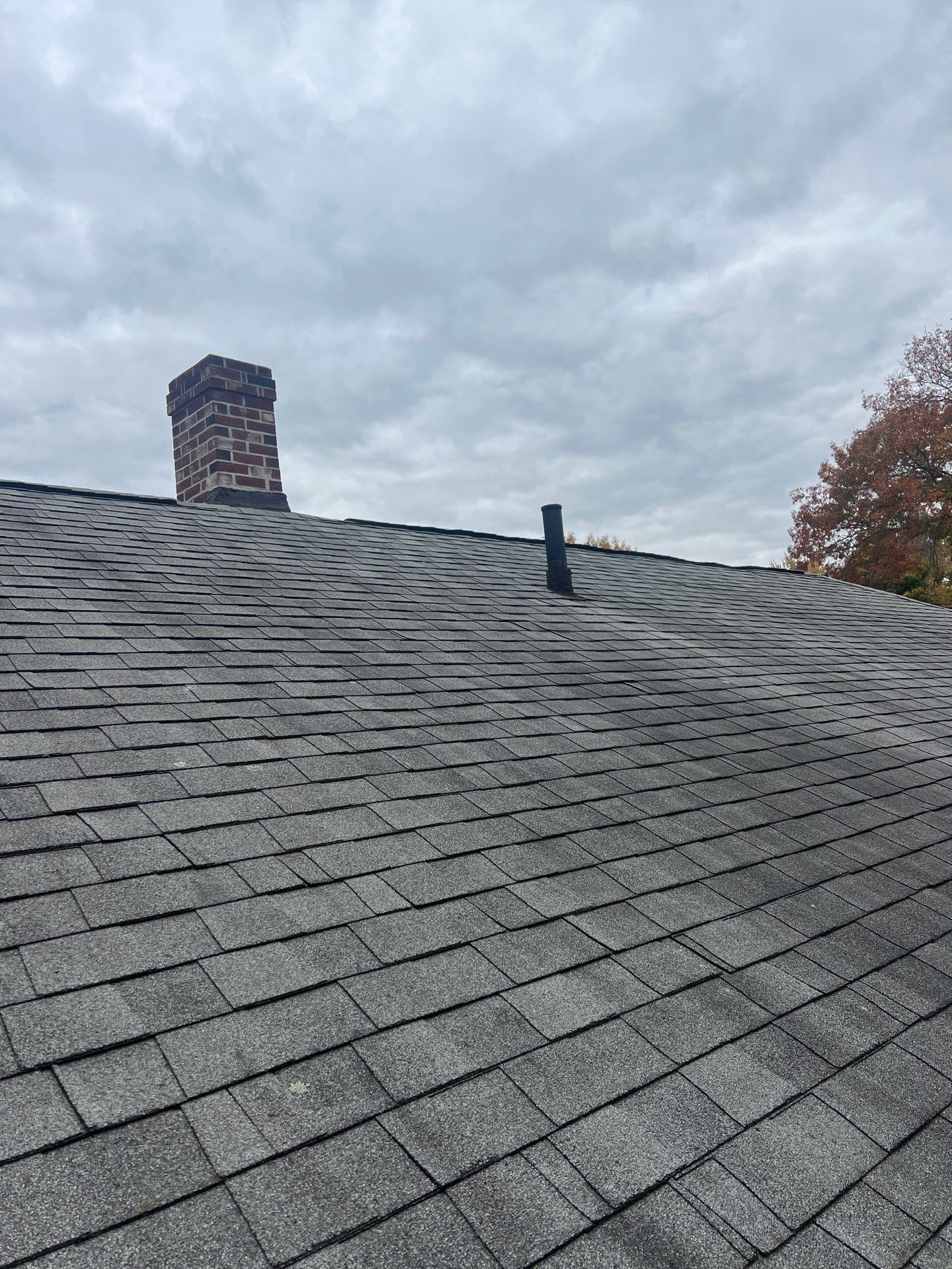 Gray asphalt shingle roof with a brick chimney and a small vent under a cloudy sky.