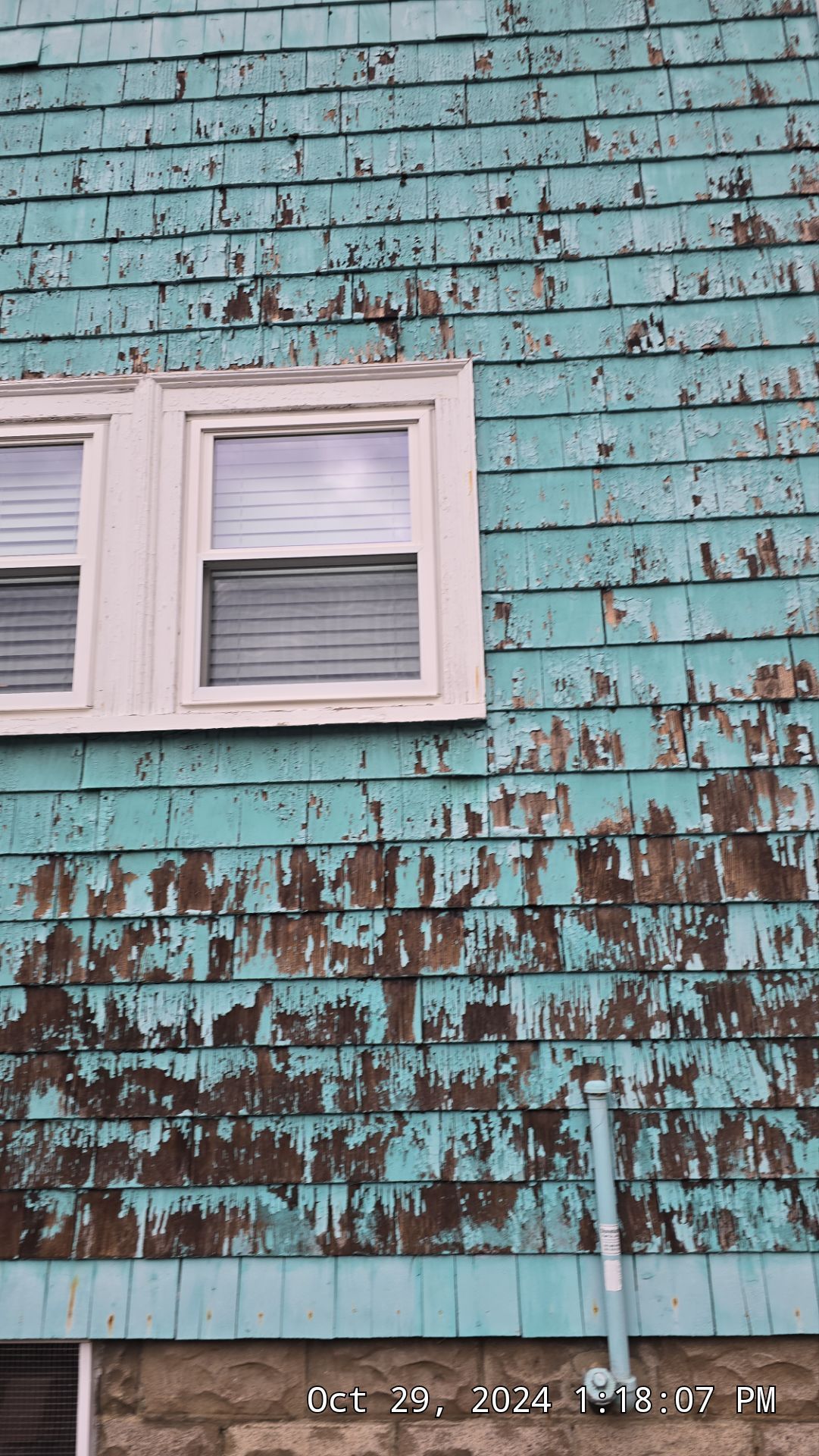 Teal shingled siding with peeling paint, a white-framed window, and a blue pipe extending down.