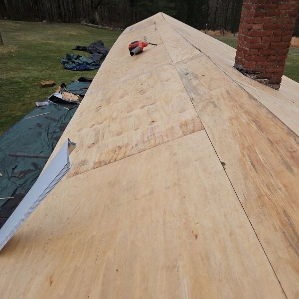 New plywood on a roof with a chimney and drill, green tarp, and trees in the background.