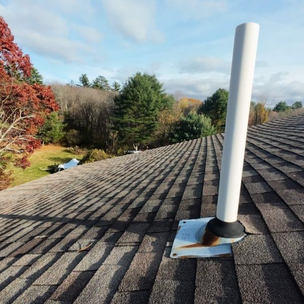 White vent pipe on a brown shingled roof with a wooded landscape background.