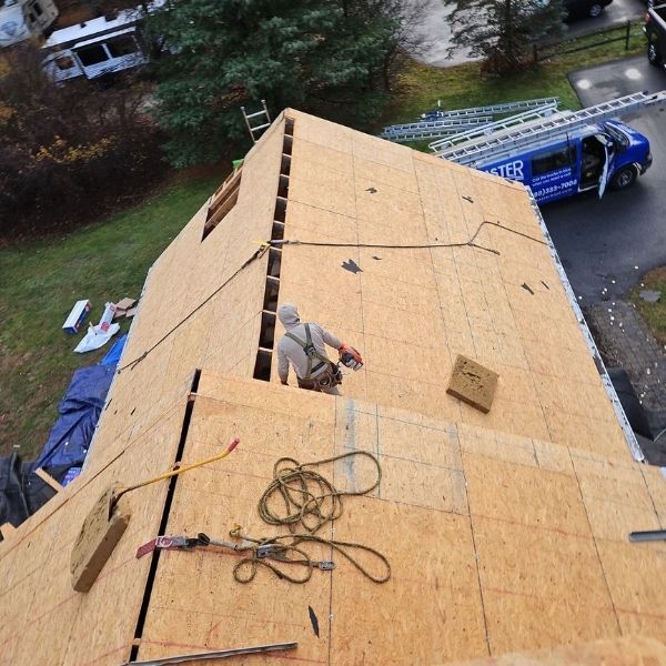 Roofer working on a house roof. Roof is covered in wood panels. Ladders and a blue truck are nearby.