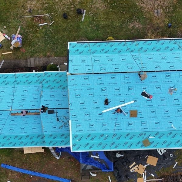 Overhead view of a roof under construction. Several workers on a blue underlayment, surrounded by tools and materials.