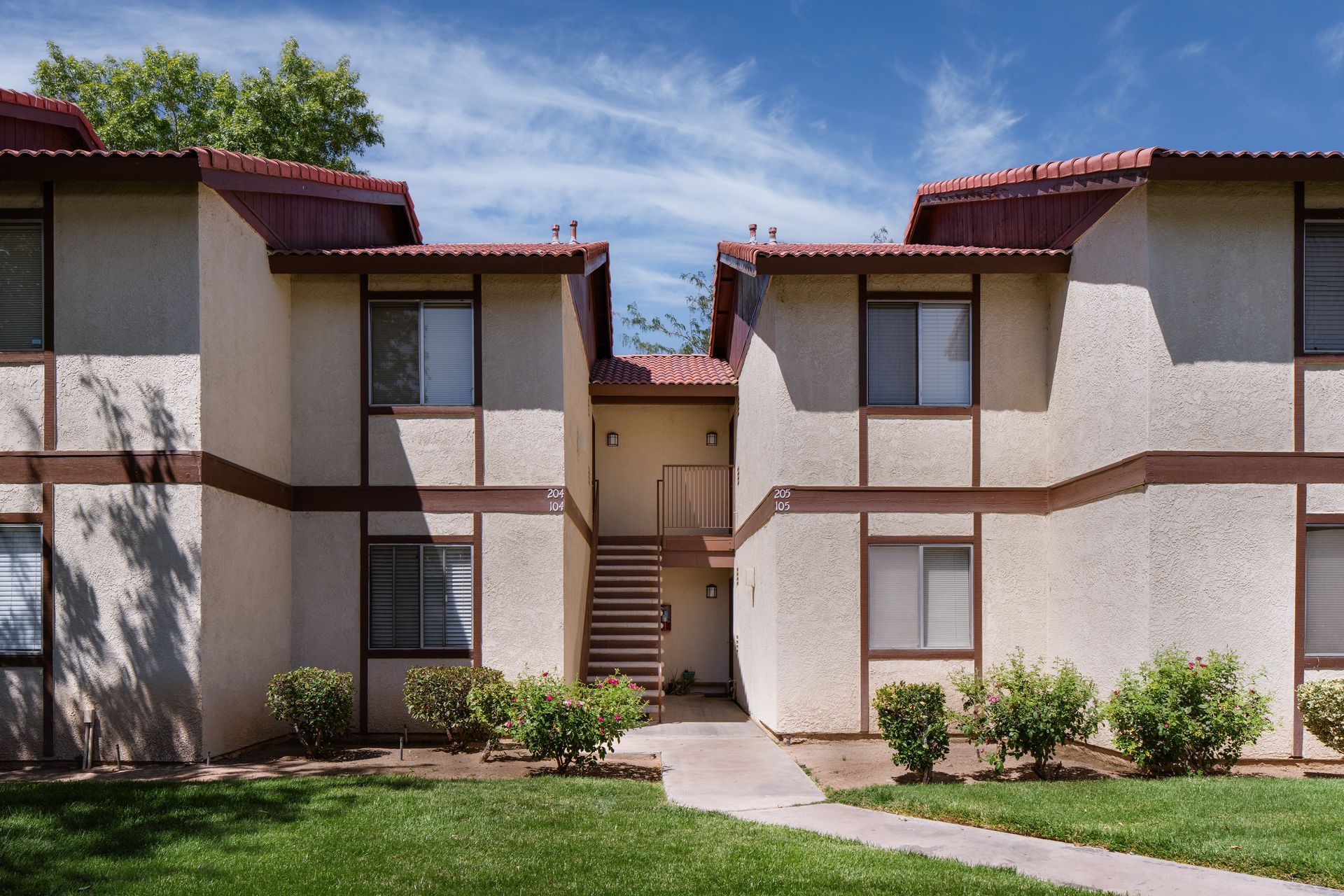 Two-story beige apartment building with brown trim and red tile roof. Green lawn and blue sky.