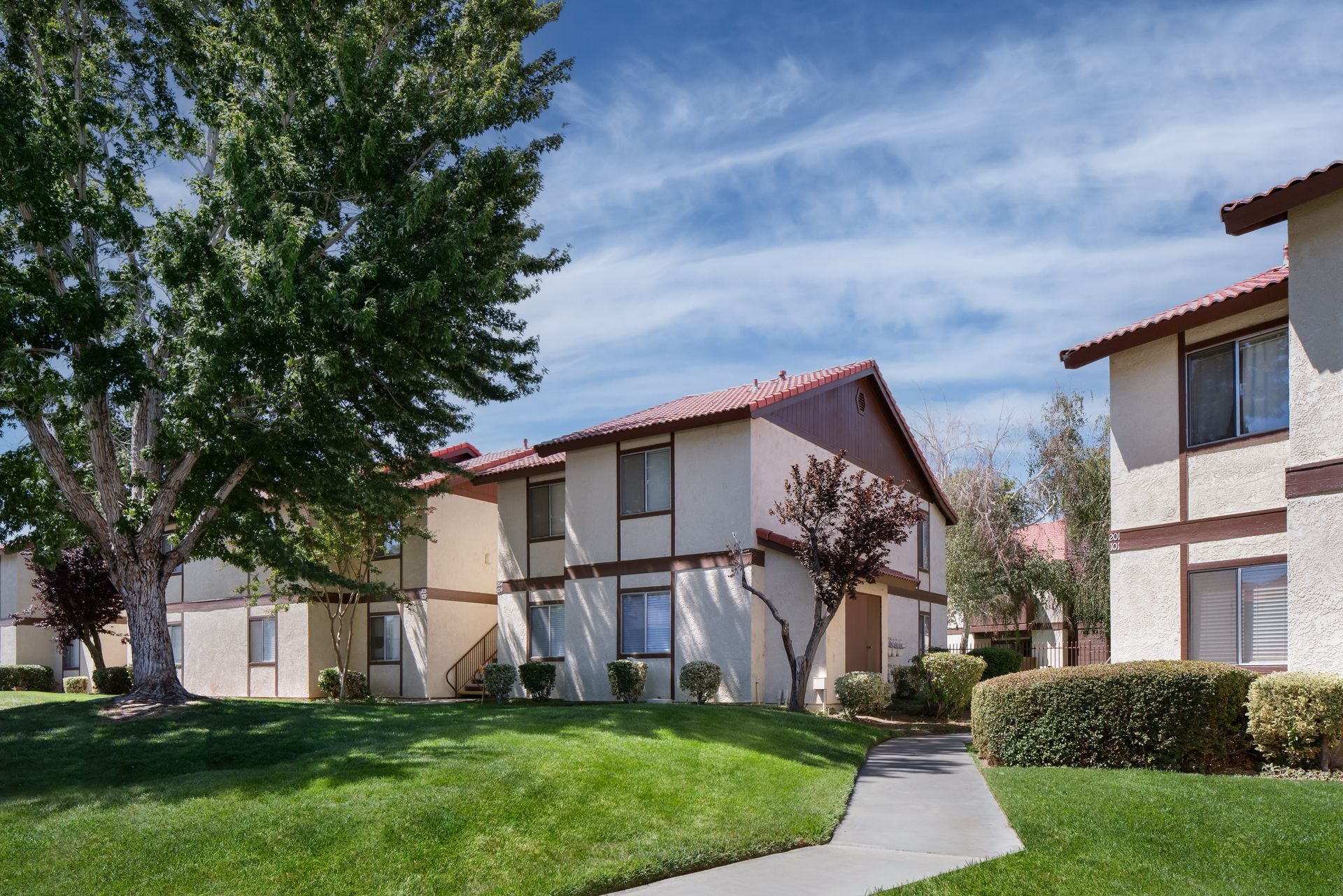 Apartment complex with beige buildings, red tile roofs, and green lawns under a blue sky.