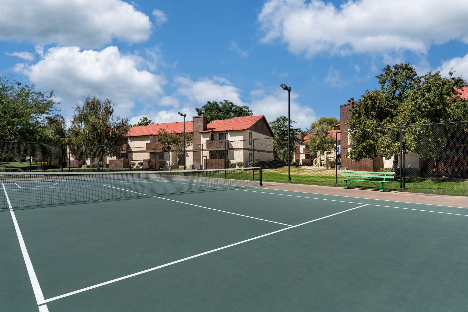 Tennis court with green surface, white lines, and apartment buildings under a blue sky.