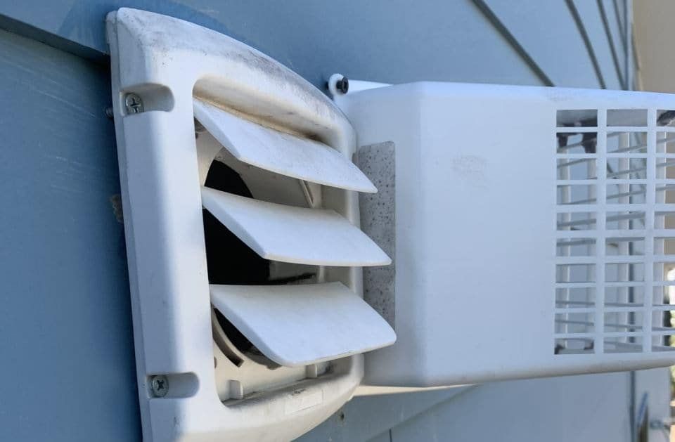 A white plastic dryer vent hood with louvers installed on the side of a blue-sided house.