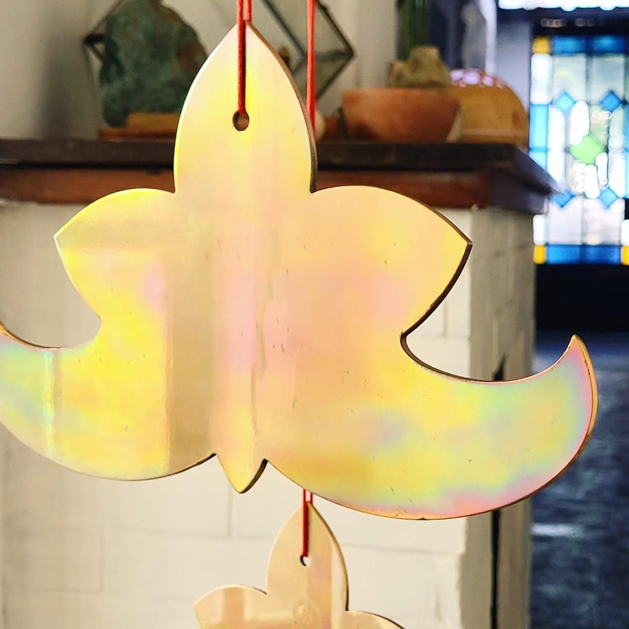 Campanas de viento doradas con forma de flor colgando en el interior; fondo borroso con plantas y una vidriera.